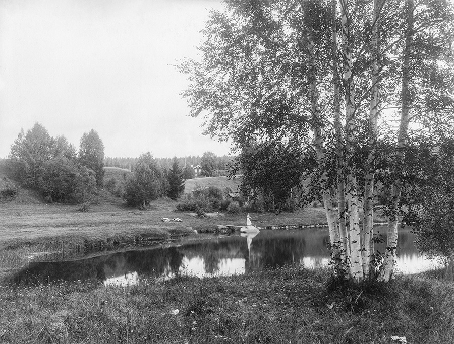 Landskap med björkar framför en sjö. I mitten av bilden står en person och metar. I bakgrunden skymtar ett hus och skog.