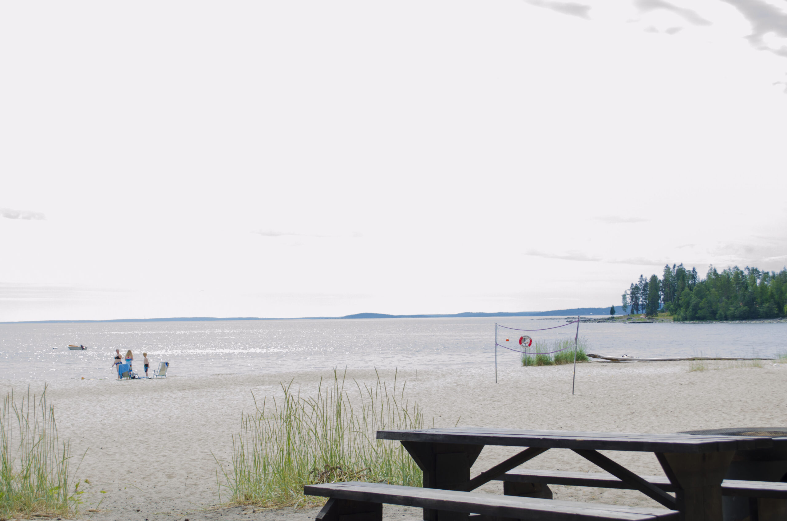 Bänkåsvikens badplats, tre badgäster på stranden på stranden finns rastmöbel och volleybollnät.