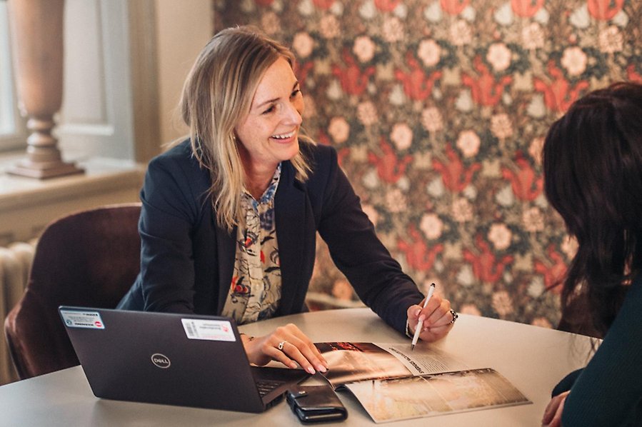 Laila Bergström sitter vid ett bord och hjälper en person med pappersarbete. Hon ser glad ut. Foto.