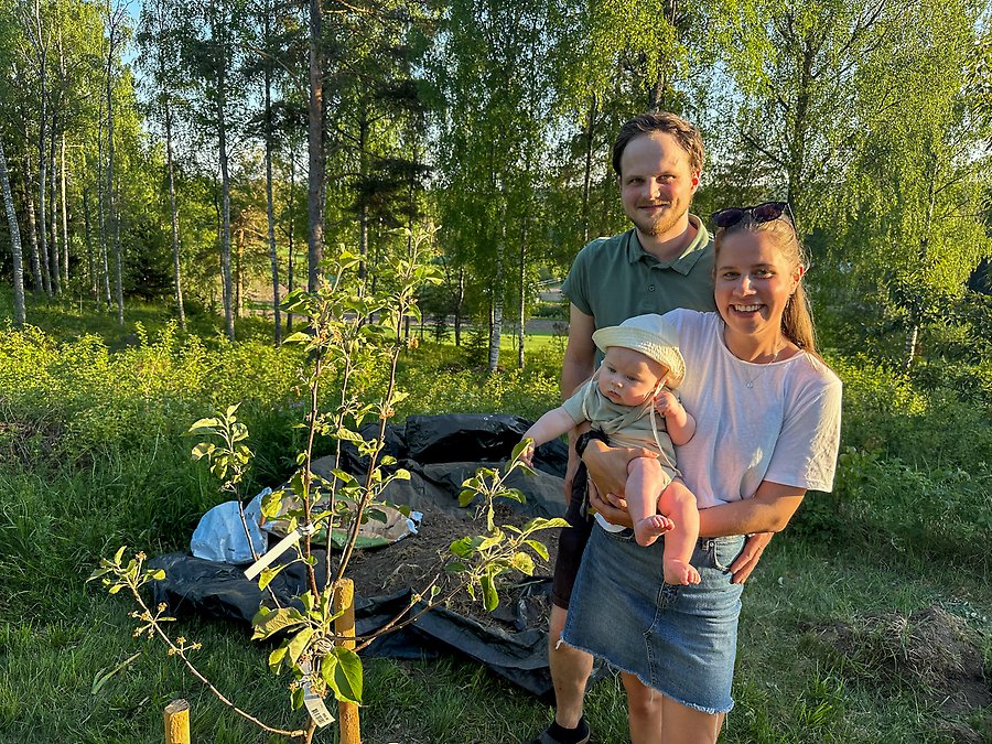 Två personer står i en trädgård. Kvinnan håller en bebis i famnen. Bredvid dem ser man ett nyplanterat träd.