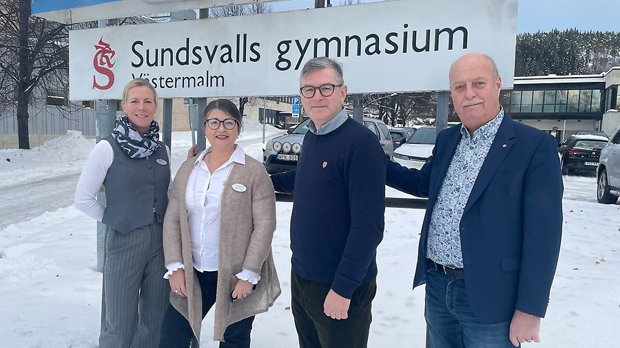 Anette Buhlér, Julia Riabouchkina, David McIntyre och Olle Åkerlund ståendes utomhus framför en stor vit skylt med svart text: Sundsvalls gymnasium. På marken ligger det mycket snö.