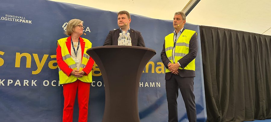 Två män och en kvinna på står på en scen med en banderoll bakom sig. På banderollen står det Sundsvalls nya logistiknav. I förgrunden syns ett svart ståbord. 