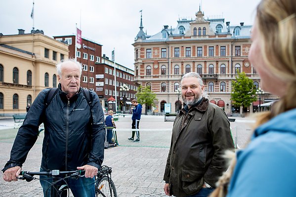 Anders Uddén står på stora torget i Sundsvall och pratar med två invånare. Bakom är Sundsvalls stenstad och människor i rörelse.