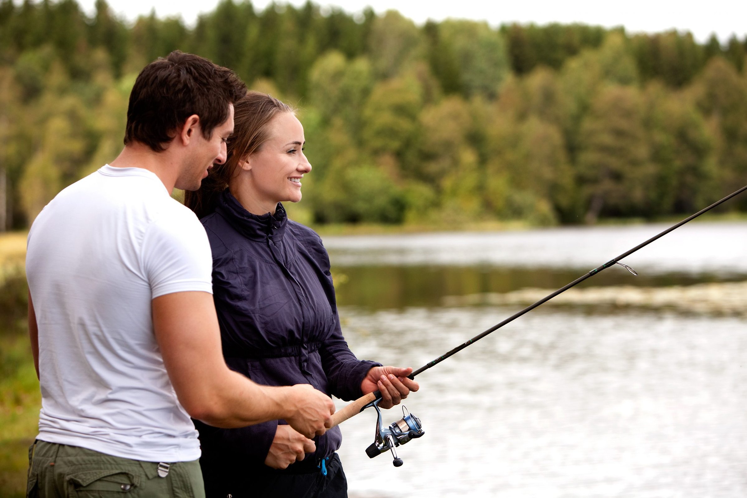 Alttext: En tjej och en kille står och håller ett fiskespö. I bakgrunden syns en sjö och skog. Fotograf: Sundsvalls kommun Bildtext: A man showing a woman how to fish